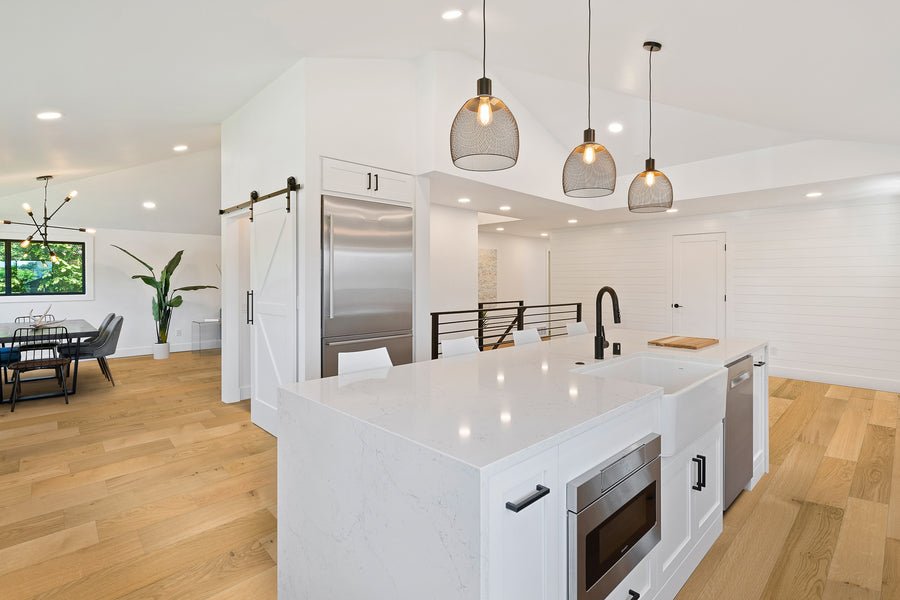 Modern kitchen with white cabinetry, marble countertops, stainless steel appliances, and pendant lights. Open layout flows into a dining area with wooden floors and black chairs. Large windows let in natural light.