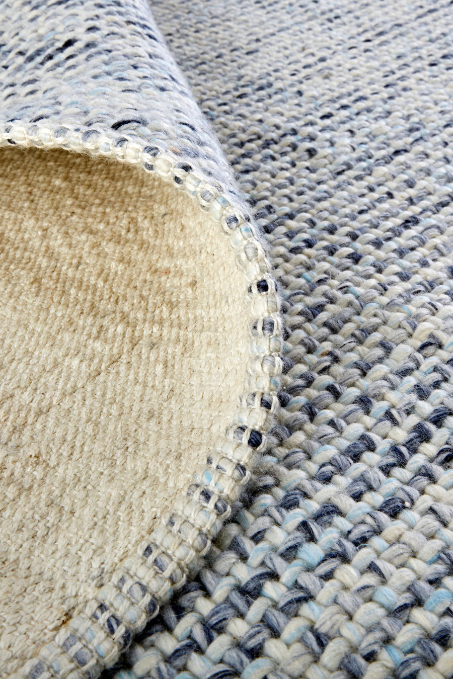 A close-up of a woven rug in neutral tones, with one corner flipped over to show the lighter, solid underside. The rug has a textured, chunky weave in shades of white, grey, and blue.