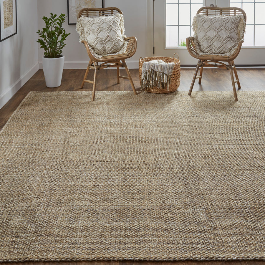 A cozy, minimal living space with a large woven jute rug, two rattan chairs with cream textured pillows, a basket with a throw blanket, a potted plant, and sunlight streaming through glass doors in the background.