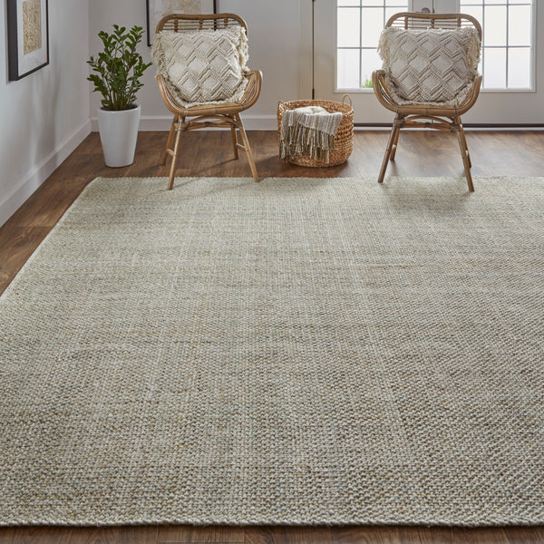 A modern living room with a large woven beige rug, two rattan chairs with textured cream cushions, a basket with a throw blanket, a potted plant, and large windows letting in natural light.