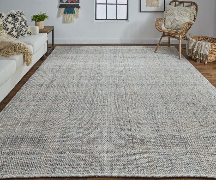 A spacious living room with a large, textured light-gray area rug on a wooden floor, a white sofa, a rattan chair with a cushion, a basket with a blanket, and modern wall decor.