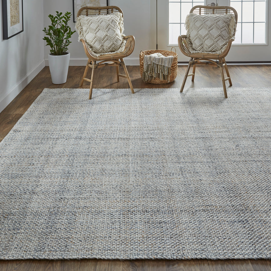 A cozy living room with a large textured gray rug, two wicker chairs with patterned cushions, a small basket with a blanket, a potted plant, wood flooring, and sunlight streaming in from glass patio doors.