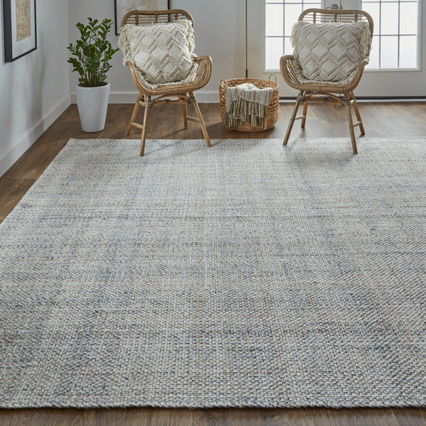 A cozy living room with a large textured gray rug, two wicker chairs with patterned cushions, a small basket with a blanket, a potted plant, wood flooring, and sunlight streaming in from glass patio doors.
