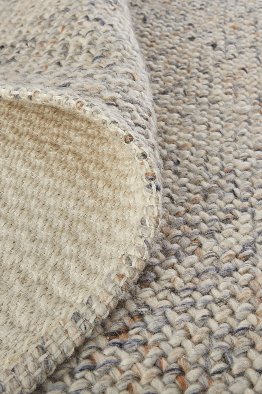 Close-up of a woven, chunky knit fabric in beige tones with specks of brown and gray. The corner is folded over, showing the texture and stitching details on both sides.