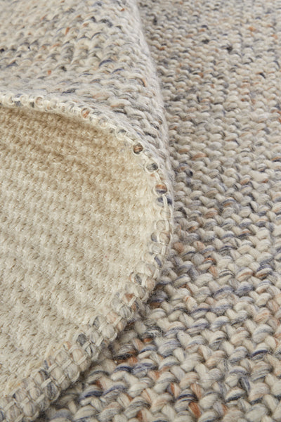 Close-up of a woven, chunky knit fabric in beige tones with specks of brown and gray. The corner is folded over, showing the texture and stitching details on both sides.