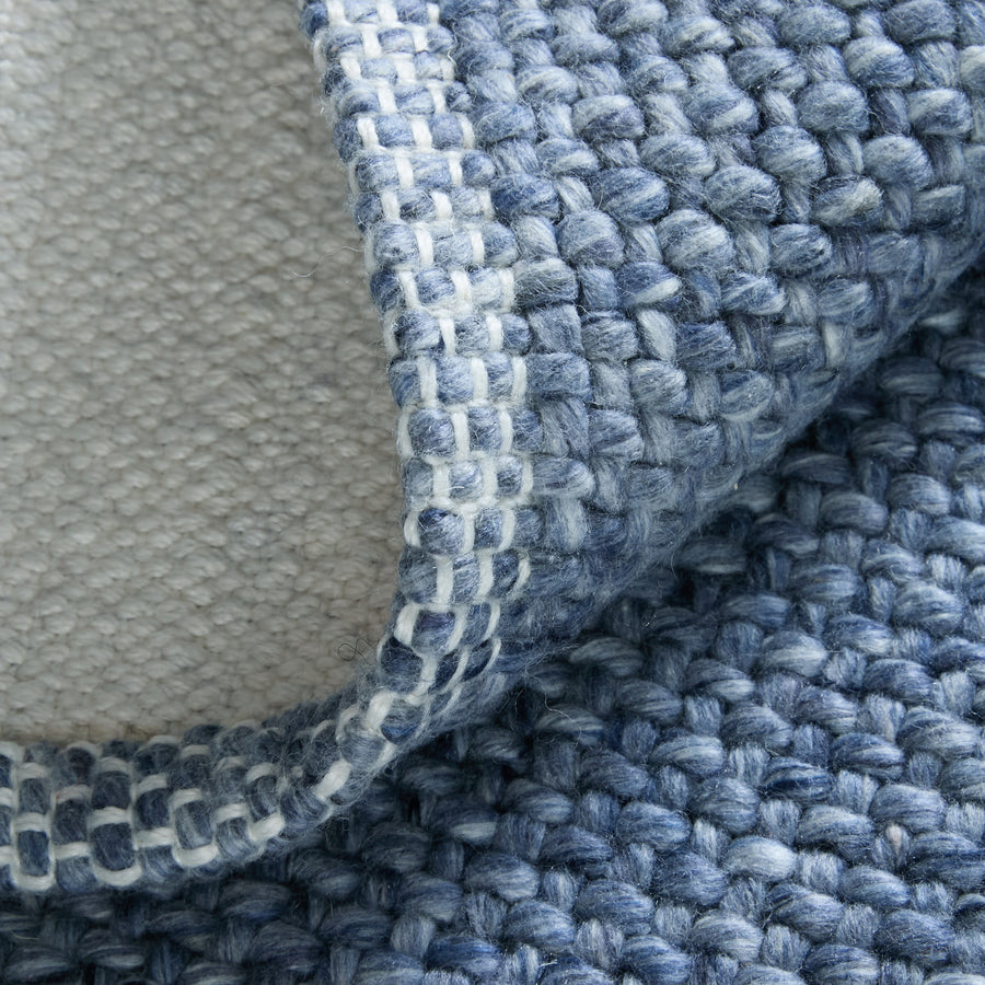 Close-up of a blue woven rug with a visible looped texture, showing the edge folded over to reveal the white and gray underside and stitched border.