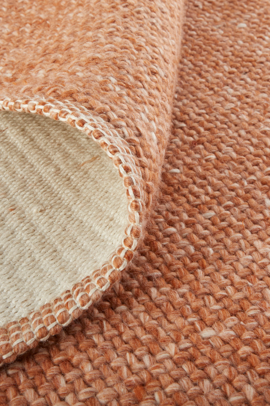 Close-up of a textured, woven area rug in shades of brown and beige, with one corner flipped over to show the lighter underside and detailed stitching along the edge.