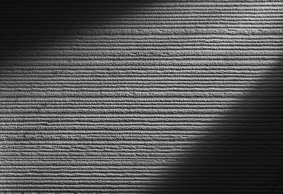 Black and white photo of a textured wall with horizontal ridges, partially illuminated by diagonal sunlight, creating a pattern of light and shadow across the surface.