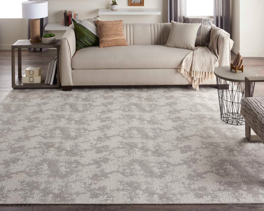 A modern living room with a beige sofa decorated with pillows and a throw, a large light gray patterned rug, a round side table, and neutral décor in a softly lit space.