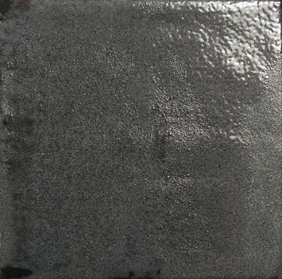 A close-up of the Nakama 5" x 5" Ceramic Tile in Graphite, showing its textured, shiny dark gray surface with uneven light reflections and a slightly rough, pebbled finish.