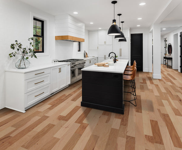 Modern kitchen with white cabinets, wood accents, a black island with bar stools, pendant lights above, and stunning Deepwood Trail 5″ Hickory Oak Hardwood flooring. A vase with greenery sits on the countertop by the window.