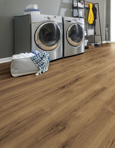 Modern laundry room with a front-loading washer and dryer, a storage basket with towels and a blanket, a gray cabinet, and wooden flooring. Shelving unit holds detergent, towels, and clothing.