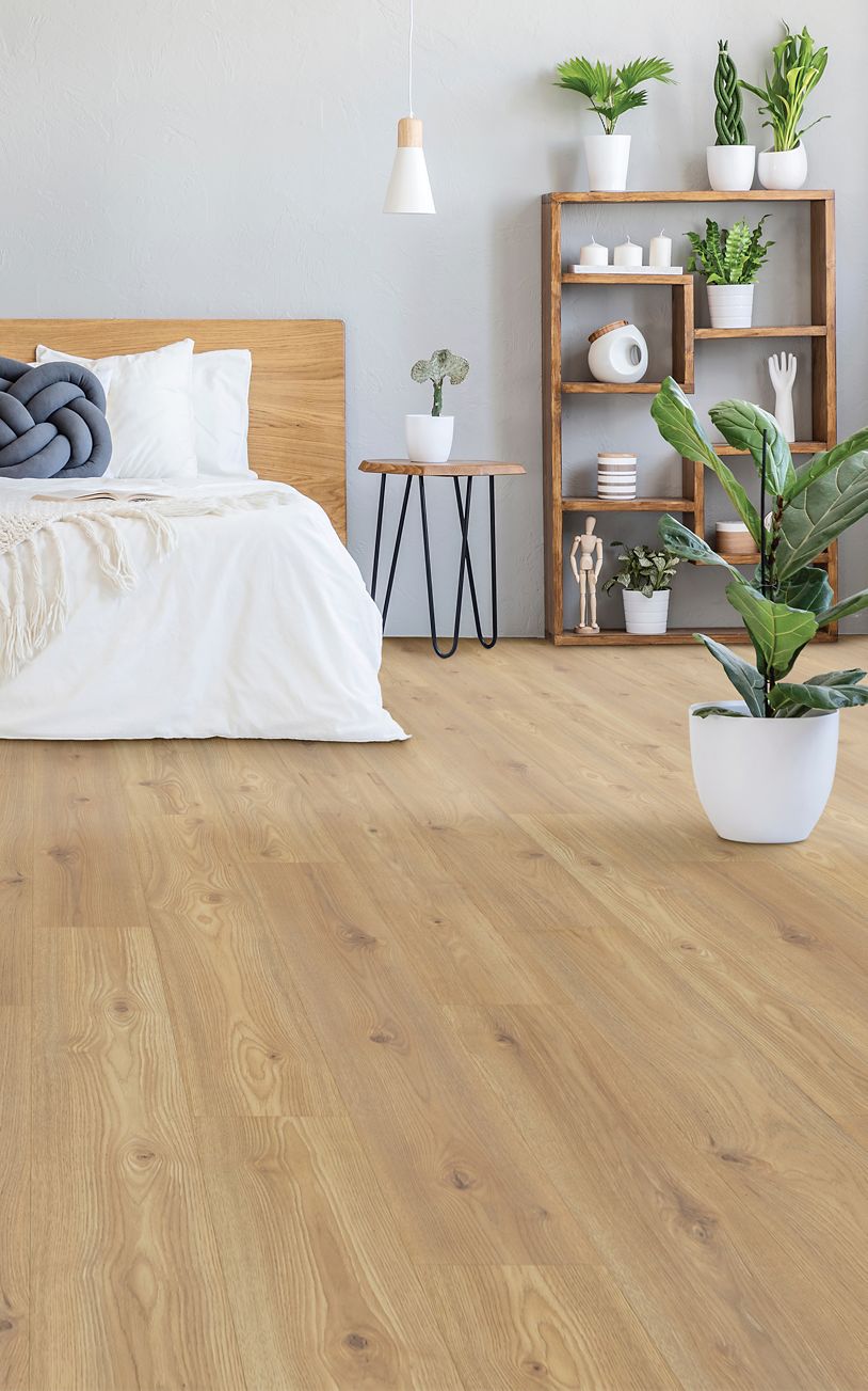 A modern bedroom featuring Freywood Springs 8 11/32" Waterproof Laminate flooring, a bed with white bedding, a wooden shelf with plants and decor, a small round side table, and a potted plant for a light, airy ambiance.