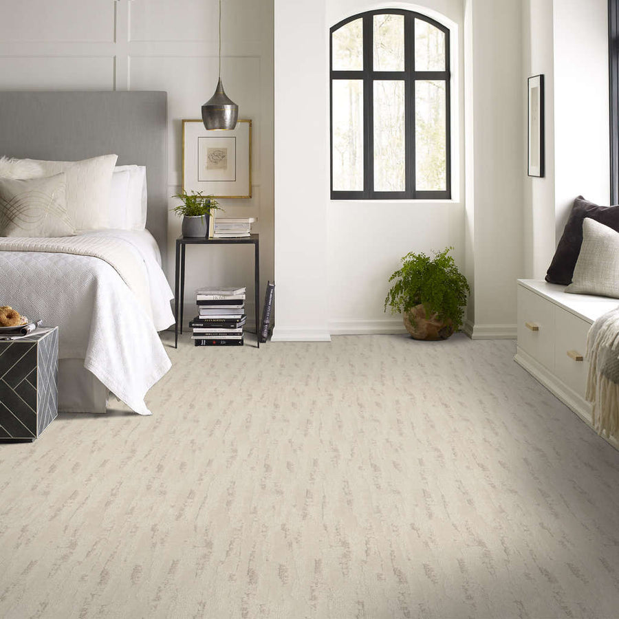 Bright, modern bedroom with a light beige carpet, a gray upholstered bed, white bedding, nightstand with lamp and decor, arched window, plant, and built-in bench with pillows along the wall.