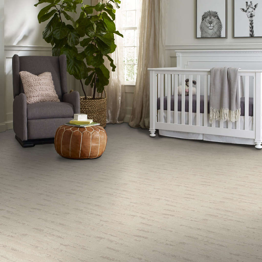 A nursery room with a gray armchair, a brown leather ottoman, a large potted plant, a white crib with a gray blanket, and animal artwork on the wall above the crib. Natural light comes in from a window.