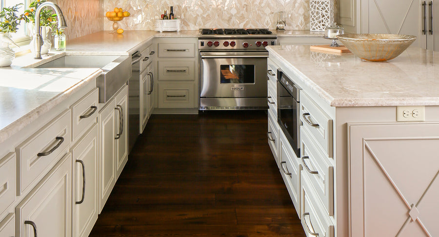Bright modern kitchen with white cabinets, marble countertops, stainless steel appliances, a farmhouse sink, a bowl on the island, and a vase of lemons by the window. Dark wood floor contrasts with the light decor.
