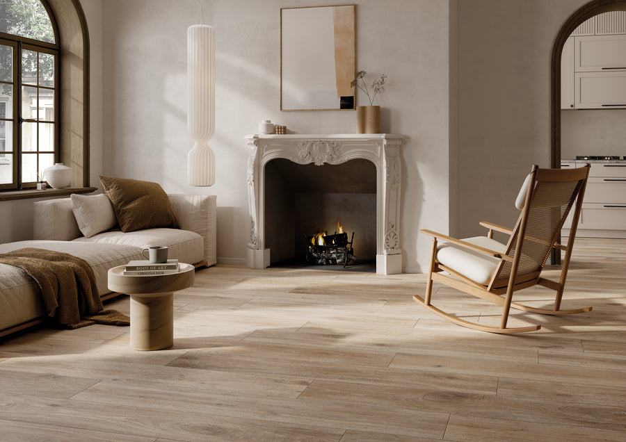 A cozy living room with light wood flooring, a white sofa with brown cushions, a modern round coffee table, a classic white fireplace with a lit fire, and a wooden rocking chair. Soft natural light streams in through the windows.