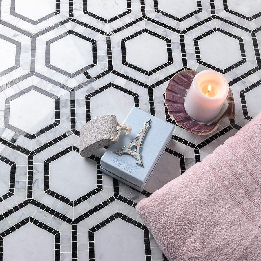 A lit candle, a folded pink towel, a pumice stone, and a book with an Eiffel Tower ornament rest on a geometric black-and-white hexagonal tile floor.