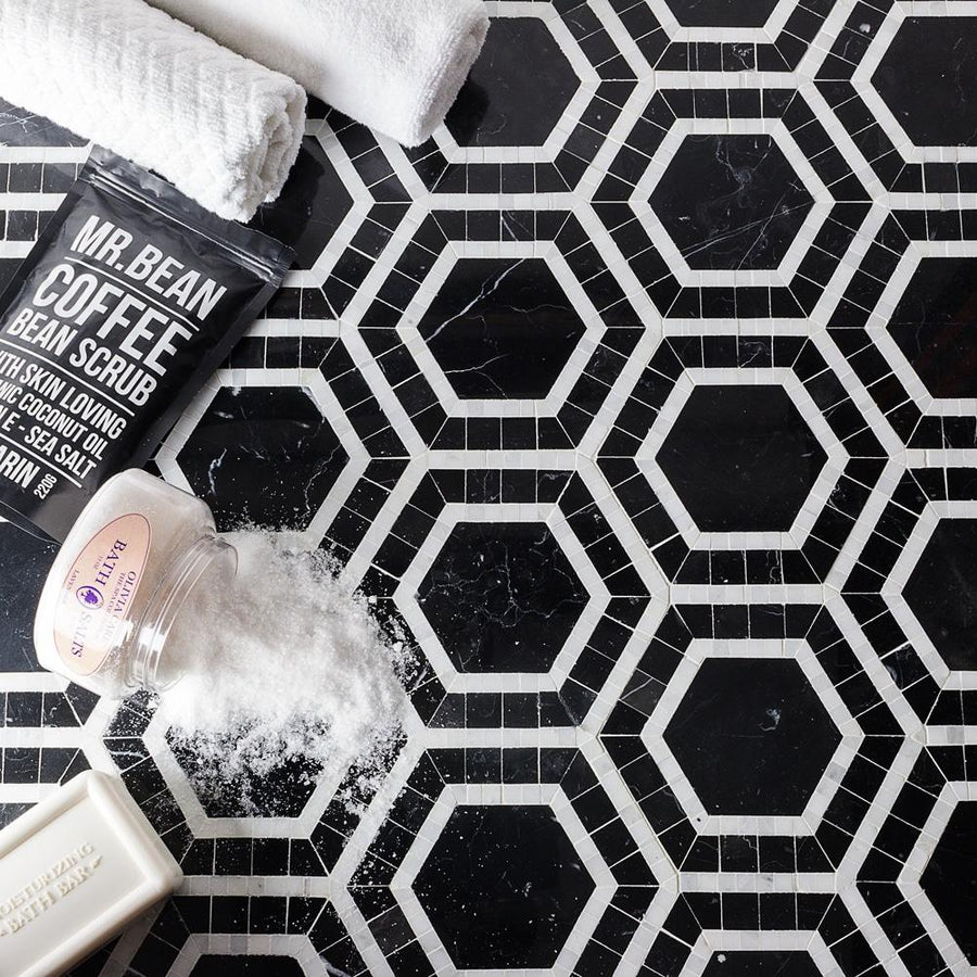 Black and white hexagon patterned tiles with a spilled jar of bath salts, a rolled white towel, a coffee bean scrub pouch, and a white soap bar arranged on the surface.