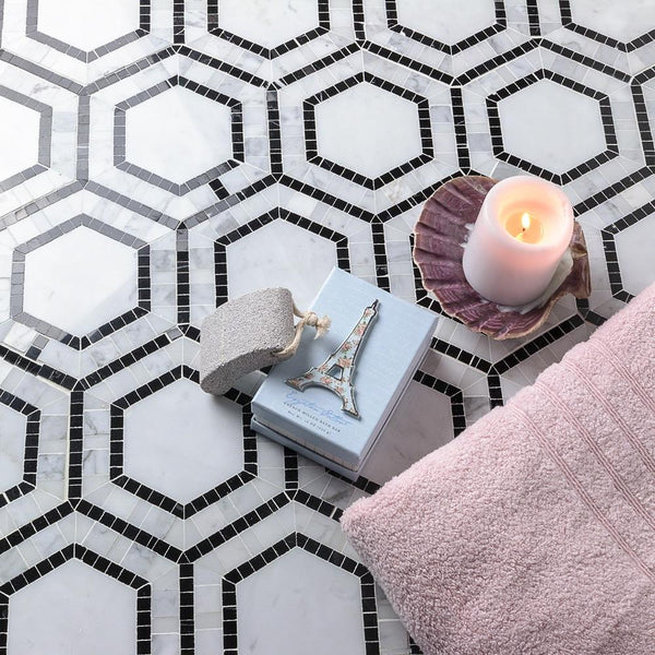 A lit candle, a folded pink towel, a pumice stone, and a book with an Eiffel Tower ornament rest on a geometric black-and-white hexagonal tile floor.