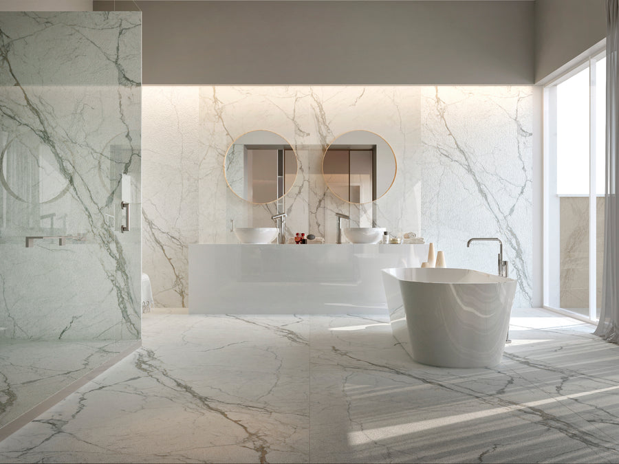 Modern bathroom with marble floors and walls, a freestanding bathtub, double sinks with round mirrors, a large glass-enclosed shower, and natural light streaming through tall windows.