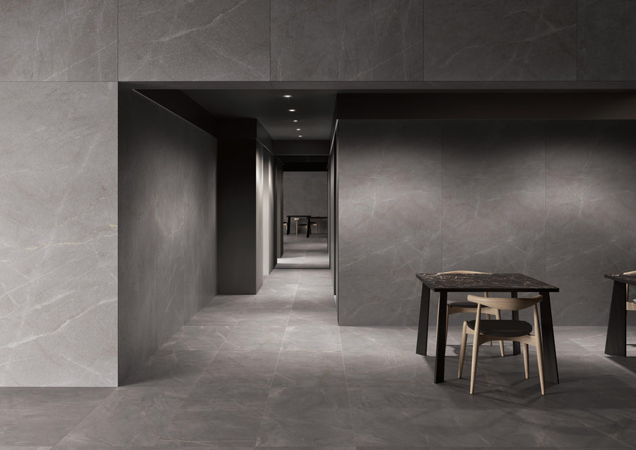 A modern interior with gray stone walls and floors, a hallway leading to dark doorways, and two dark tables each paired with a light wooden chair in the foreground.
