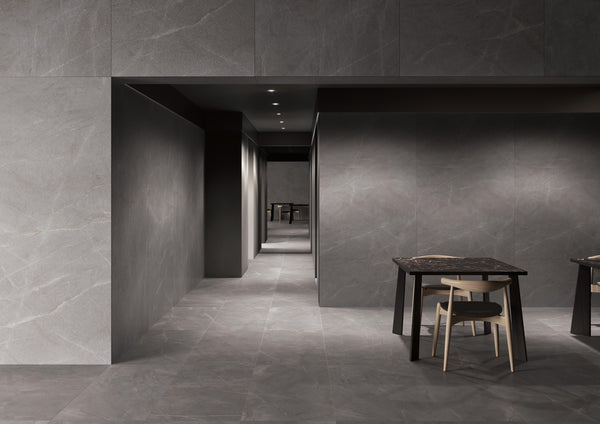 A modern interior with gray stone walls and floors, a hallway leading to dark doorways, and two dark tables each paired with a light wooden chair in the foreground.