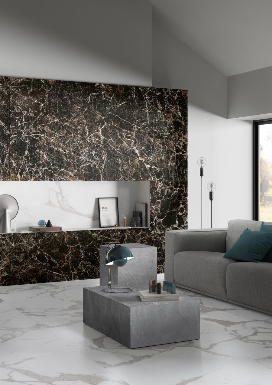 A modern living room with a grey sofa, blue cushions, a square stone coffee table, and marble-patterned floors. The wall behind features dark marble with white veins, and there are minimalist decor items and floor lamps by a large window.