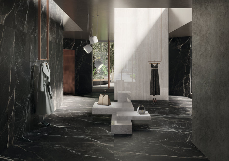 A modern, minimalist clothing store with dark marble walls and floors, hanging garments, geometric white display tables, and sunlight streaming through tall windows onto stylish accessories.