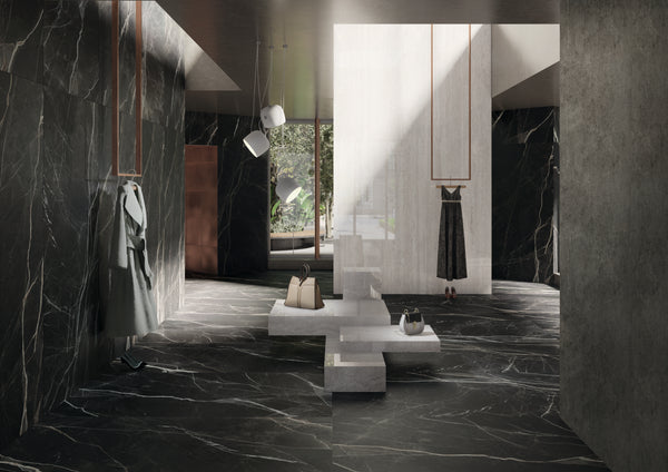 Minimalist, modern interior with dark marble walls and floors, geometric white seating, hanging lights, and clothing items on display. Sunlight streams in from a tall window, casting soft shadows in the stylish space.