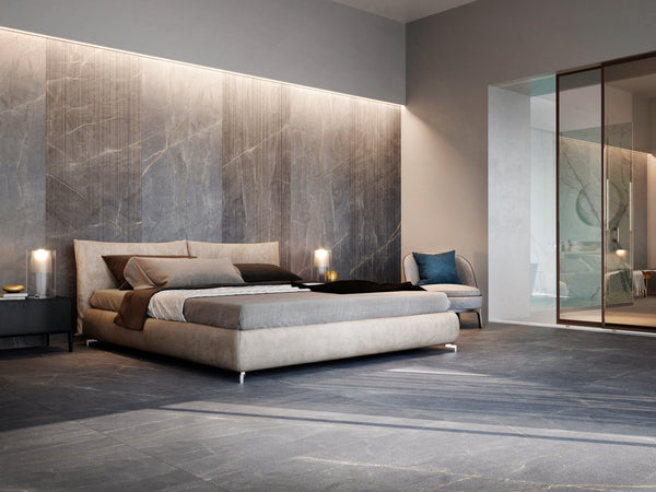 Modern bedroom features a large beige bed, dark accent bedding, bedside tables with lamps, a blue-gray chair, and floor-to-ceiling Donatello 24x48 3D Rectified Porcelain Tile in gray. A glass wall reveals the bathroom area.
