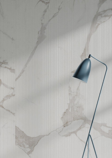 A modern black floor lamp stands in front of a white wall with a subtle marble pattern and vertical textured lines, illuminated by natural light casting soft shadows.