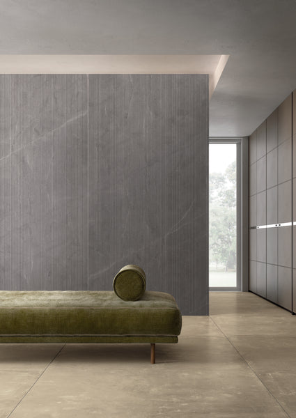 A minimalist interior featuring a green upholstered bench with a round cushion, light beige flooring, a textured gray accent wall, and a large window letting in natural light.