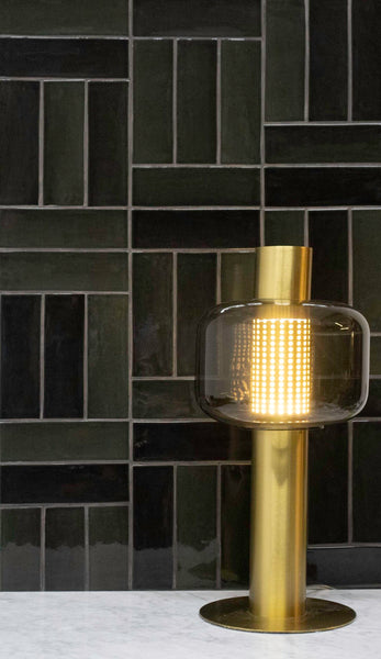 A modern brass table lamp with a cylindrical glass shade sits on a white marble surface in front of a dark green tiled wall with a geometric pattern.