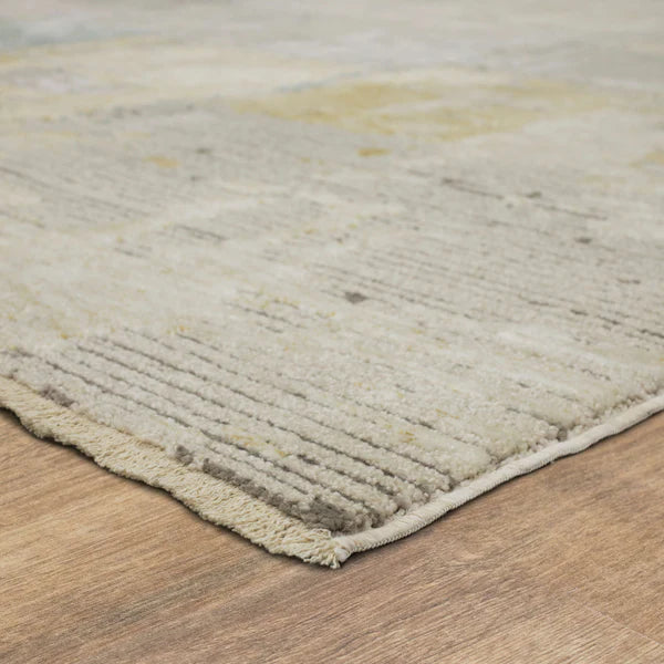Close-up view of a light-colored, textured area rug with subtle gray and beige tones, placed on a wooden floor. The rug has a soft, slightly uneven surface and visible woven edges.