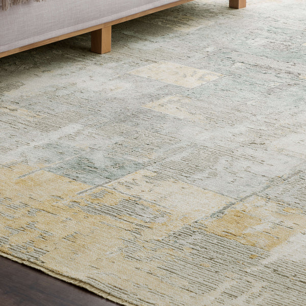 A close-up view of a light-colored area rug with a subtle, abstract pattern in beige, yellow, and pale green tones, placed near the wooden legs of a beige sofa on a dark wood floor.