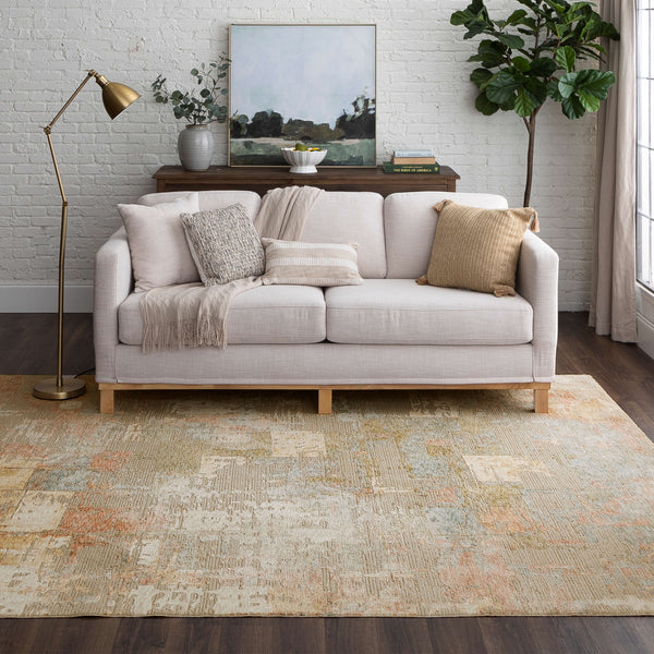 A cozy living room with a light beige sofa, neutral throw pillows, a knit blanket, a brass floor lamp, a wooden console table with decor, a leafy plant, and a large painted rug on dark wood floors.