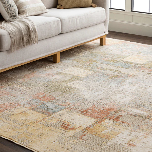 A beige and pastel area rug with an abstract pattern lies on a dark wood floor in front of a light-colored, modern sofa with tan pillows and a beige throw, next to a window with white trim.