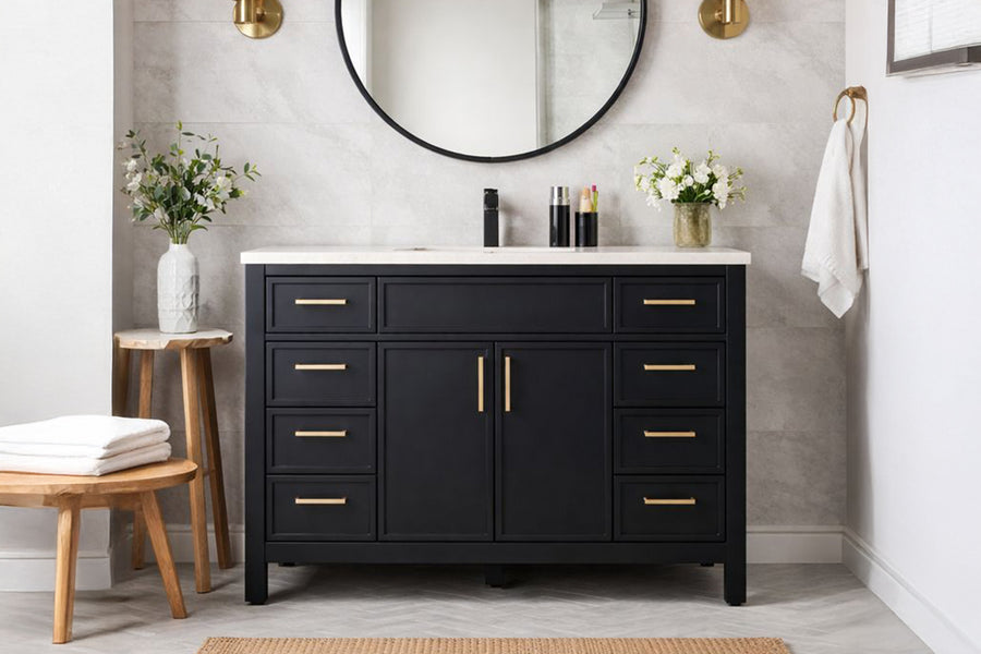 A modern bathroom with the Novello 48" Vanity in black and gold, a white countertop, round mirror, wall sconces, small plants on each side; towels rest on a wooden stool and another towel hangs nearby.