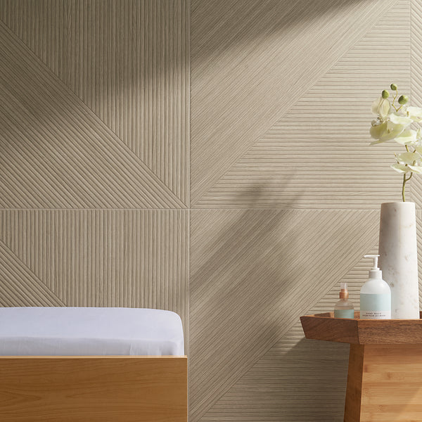A minimalist spa setting with geometric beige wall tiles, a wooden bench with a white cushion, and a wooden table holding a white vase with flowers and bottles of soap and lotion.
