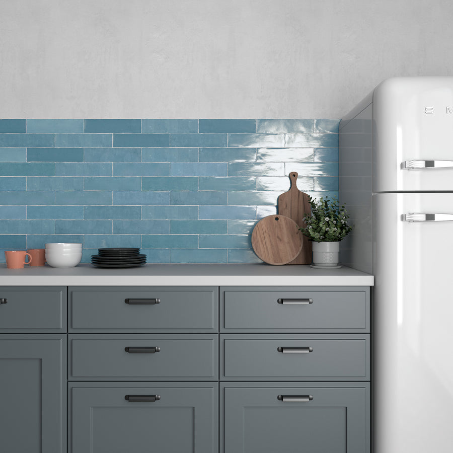 A modern kitchen with blue cabinets, a white countertop, stacked dishes, a cutting board, a potted plant, and a white retro-style refrigerator against a wall of glossy blue subway tiles.