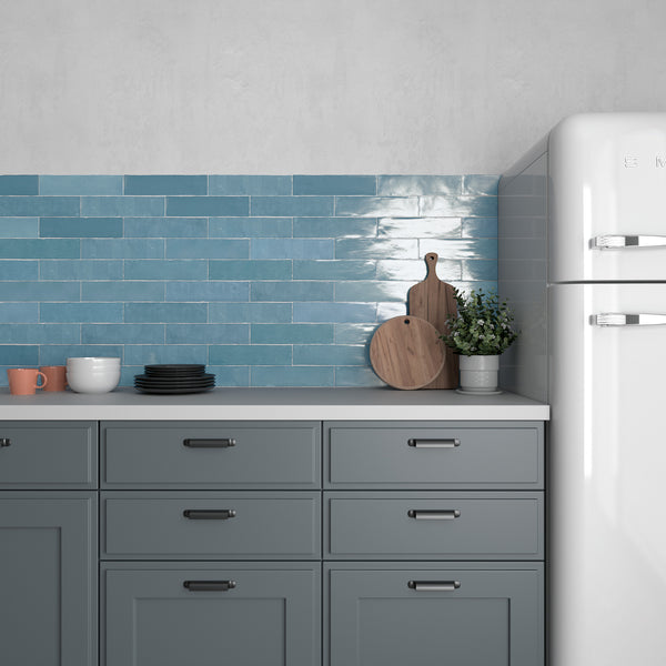 A modern kitchen with blue cabinets, a white countertop, stacked dishes, a cutting board, a potted plant, and a white retro-style refrigerator against a wall of glossy blue subway tiles.