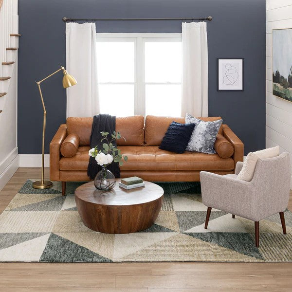 A modern living room with a tan leather sofa, gray accent chair, round wooden coffee table, geometric rug, floor lamp, and white curtains over a large window. Cushions and decor add a cozy touch.