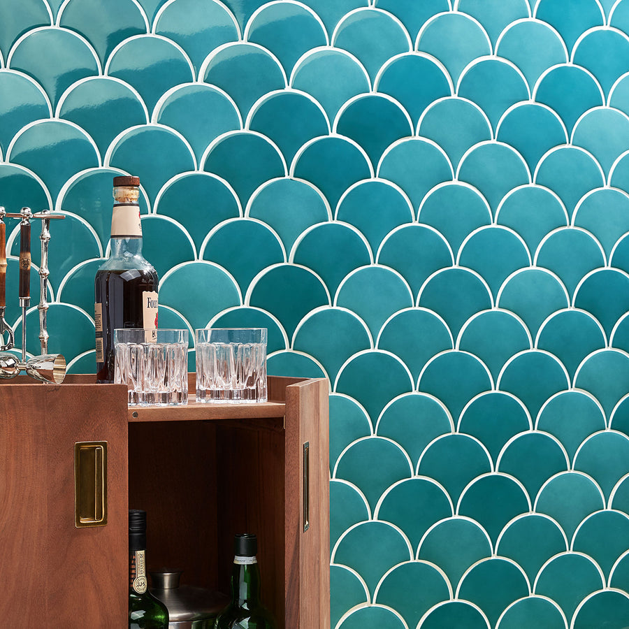 A wooden bar cabinet with bottles and glasses in front of a teal fish scale tile wall with white grout. The cabinet is partially open, showing more bottles inside.