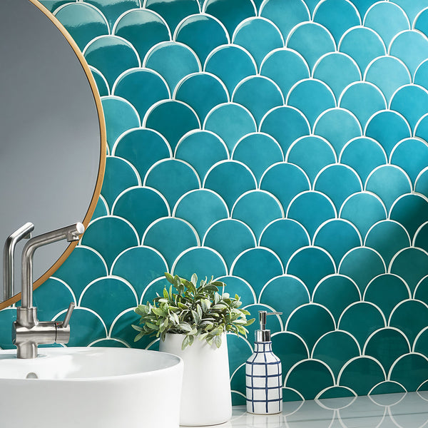 A bathroom sink with a silver faucet, a potted plant, and a soap dispenser sits in front of a teal fish scale tile backsplash and a round mirror with a gold rim.