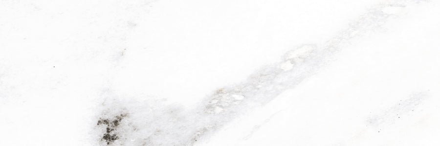 White marble surface with subtle gray veining and a few darker gray spots. The overall appearance is smooth, with natural patterns giving the stone a simple, elegant texture.