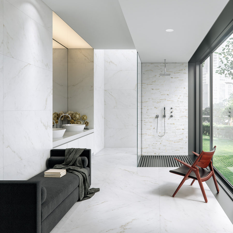 Modern bathroom with white marble walls and floor, a walk-in shower with glass partition, a black sofa with a blanket and books, a wooden chair, and large windows overlooking a green lawn.