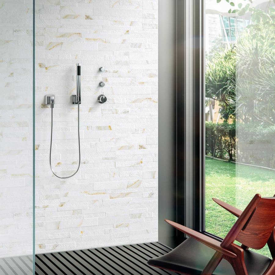 A modern shower with a handheld showerhead on a white textured tile wall, next to large glass windows overlooking a green garden. A wooden chair sits nearby on dark slatted flooring.