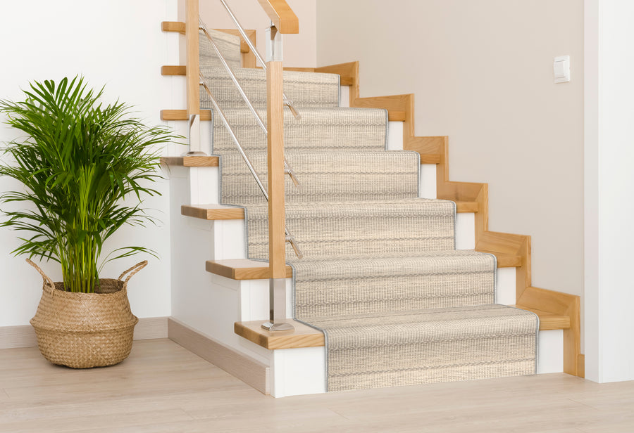 A light-colored, carpeted staircase with wooden handrails and metal balusters is next to a large green plant in a woven basket, set in a bright, modern interior with light walls and flooring.