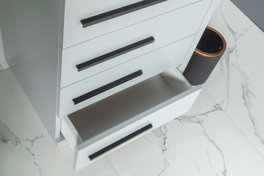 A white cabinet with four drawers, one of which is open, is positioned on a white marble floor with dark veins. A black cylindrical trash bin sits beside the cabinet.